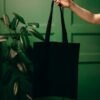 Minimalist black tote bag held by a hand against a lush green background with a leafy plant.